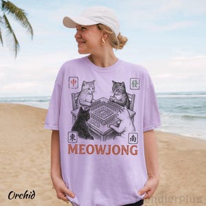 Może przedstawiać: Jasnofioletowy t-shirt z grafiką przedstawiającą cztery koty siedzące przy stole i grające w mahjonga. Wz&oacute;r zawiera chińskie znaki w czterech rogach oraz napis MEOWJONG wydrukowany pogrubioną, pomarańczową czcionką pod stołem.