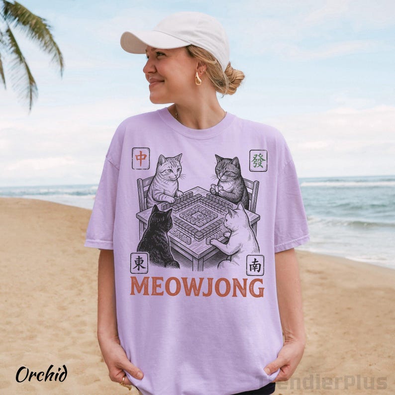 Może przedstawiać: Jasnofioletowy t-shirt z grafiką przedstawiającą cztery koty siedzące przy stole i grające w mahjonga. Wz&oacute;r zawiera chińskie znaki w czterech rogach oraz napis MEOWJONG wydrukowany pogrubioną, pomarańczową czcionką pod stołem.