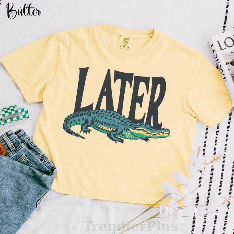 May include: A pale yellow short-sleeve t-shirt featuring the word LATER printed in large dark gray capital letters above a graphic illustration of a green and teal alligator. The shirt has a crew neckline and a standard fit.