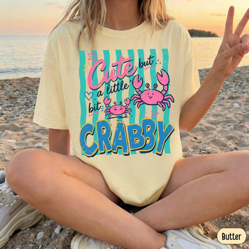 May include: A pale yellow short-sleeve t-shirt featuring a graphic print of two pink crabs and teal vertical stripes. The text reads Cute but a little bit CRABBY in a mix of pink script and bold blue block lettering with small star accents.
