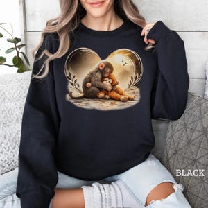 Puede incluir: Una sudadera negra de manga larga con un gr&aacute;fico central de un mono abrazando a un peque&ntilde;o juguete de peluche de mono. El dise&ntilde;o est&aacute; enmarcado por un contorno dorado en forma de coraz&oacute;n con hojas y un fondo suave y brillante.