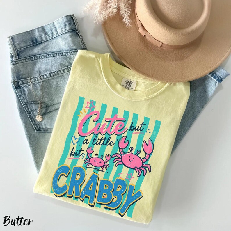 May include: A pale yellow t-shirt featuring a graphic print with teal vertical stripes, two pink crabs, and the text "Cute but a little bit CRABBY" in pink and blue lettering. The shirt is paired with light blue denim jeans and a tan wide-brim hat.