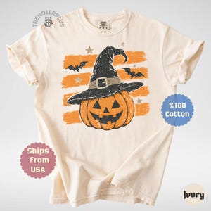 Puede incluir: Una camiseta de manga corta color marfil con un gr&aacute;fico de una calabaza de Halloween que lleva un sombrero de bruja negro. El dise&ntilde;o incluye trazos de pincel horizontales de color naranja, murci&eacute;lagos negros y peque&ntilde;as estrellas.