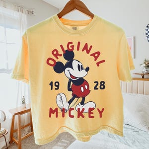 May include: A pale yellow short-sleeve t-shirt featuring a graphic of Mickey Mouse in the center. The text "ORIGINAL" is arched above the character, "19" and "28" are on the sides, and "MICKEY" is printed in red below the character illustration.