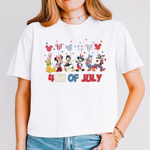 May include: A white short-sleeved t-shirt featuring a graphic of six cartoon characters holding balloons shaped like Mickey Mouse ears. The characters are dressed in red, white, and blue patriotic attire. Below the graphic is the text 4TH OF JULY.