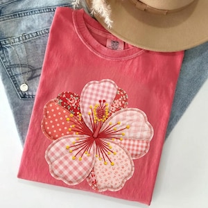 May include: A coral-colored t-shirt featuring a large, appliqued flower design. The flower is made of various patterned fabrics, including polka dots, gingham, and floral prints. A tan hat and blue jeans are also in the image.