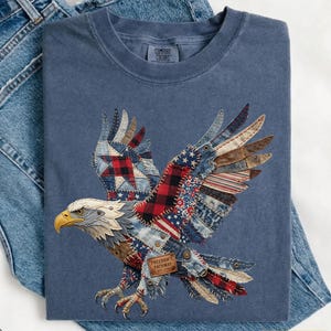 May include: A blue Comfort Colors t-shirt featuring a patchwork eagle design. The eagle's wings incorporate red, white, and blue patterns, including stars and stripes. The text "Freedom's Pathway Est. 1776" is on a small tag. The word "Denim" is in the bottom left corner.