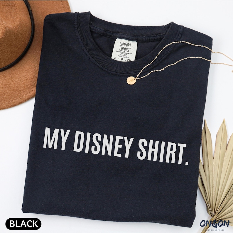 May include: A black t-shirt with the text "MY DISNEY SHIRT." in white block letters. A gold necklace with a small pendant is draped across the shirt. The shirt has a Comfort Colors tag.