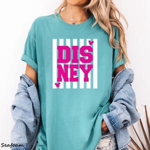 May include: Seafoam green t-shirt with the word "DISNEY" in bright pink block letters. The word is set against a white striped background. Small pink Mickey Mouse silhouettes are on the top and bottom of the word. The shirt is a casual style.