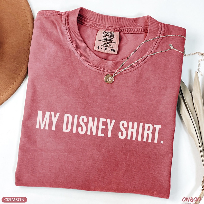 May include: Crimson-colored t-shirt with the text "MY DISNEY SHIRT." A silver necklace with a pendant rests on the shirt. The shirt is folded and has a Comfort Colors tag.