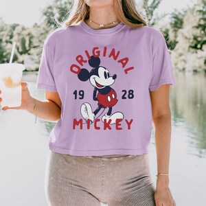 May include: A light purple short-sleeve t-shirt featuring a graphic of Mickey Mouse in the center. The text reads ORIGINAL in a red arc above the character, 19 and 28 on either side, and MICKEY in red letters below the character's white shoes.