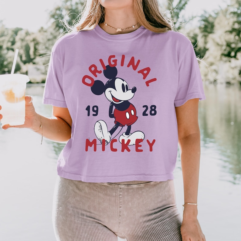 May include: A light purple short-sleeve t-shirt featuring a graphic of Mickey Mouse in the center. The text reads ORIGINAL in a red arc above the character, 19 and 28 on either side, and MICKEY in red letters below the character's white shoes.