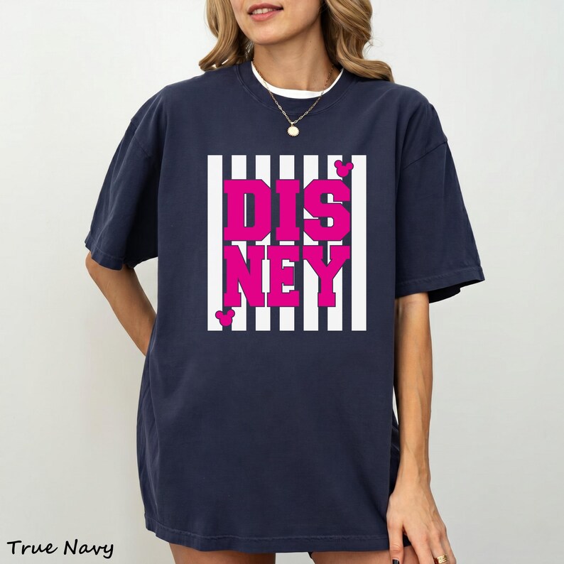 May include: Navy blue t-shirt with the word "DISNEY" in bright pink letters, set against a white and black striped background. The shirt has a classic crew neck and short sleeves. The text "True Navy" is at the bottom left.