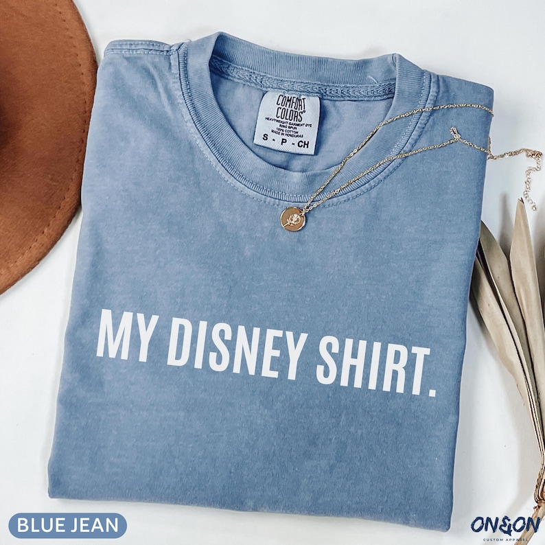 May include: A blue jean colored t-shirt with the text "MY DISNEY SHIRT." in white block letters. A gold necklace with a pendant rests on the shirt. The shirt is folded and has a Comfort Colors tag.