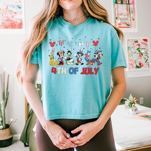 May include: A light blue short-sleeve t-shirt featuring a graphic of Disney characters including Pluto, Minnie Mouse, Donald Duck, Mickey Mouse, Daisy Duck, and Goofy holding balloons. The text 4TH OF JULY is printed in red and blue below the characters.