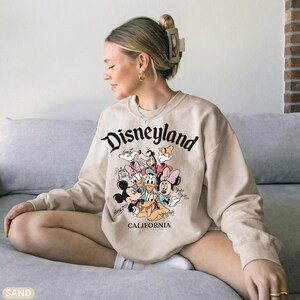 May include: Beige sweatshirt with the word "Disneyland" in black script. The sweatshirt features a colorful graphic of Disney characters, including Mickey Mouse, Minnie Mouse, and Donald Duck. The word "CALIFORNIA" is printed below the graphic.