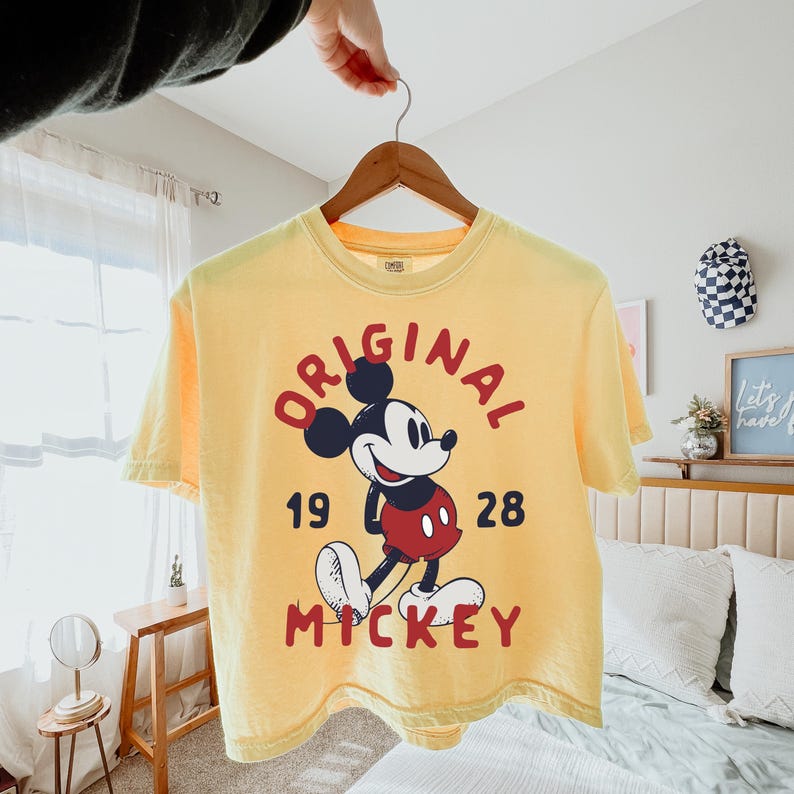 May include: A pale yellow short-sleeve t-shirt featuring a graphic of Mickey Mouse in the center. The text "ORIGINAL" is arched above the character, "19" and "28" are on the sides, and "MICKEY" is printed in red below the character illustration.