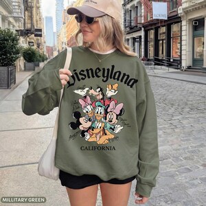 May include: Military green sweatshirt with the word "Disneyland" in black script. The sweatshirt features a graphic of Disney characters including Mickey Mouse, Minnie Mouse, Donald Duck, and Goofy. The word "California" is printed below the graphic.