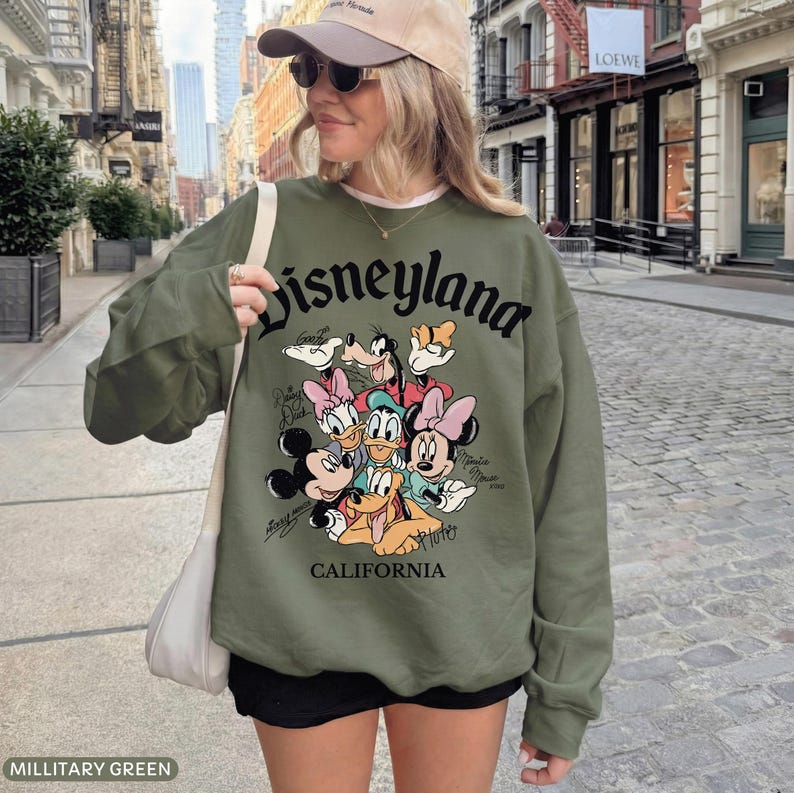May include: Military green sweatshirt with the word "Disneyland" in black script. The sweatshirt features a graphic of Disney characters including Mickey Mouse, Minnie Mouse, Donald Duck, and Goofy. The word "California" is printed below the graphic.
