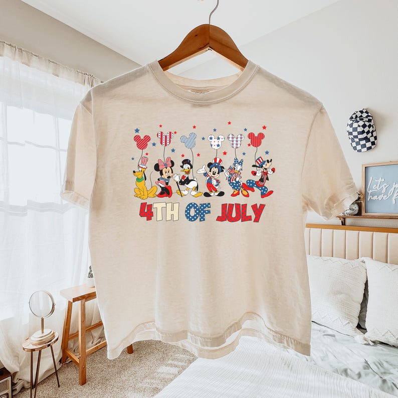 May include: A cream-colored short-sleeve t-shirt featuring a graphic of six cartoon characters holding Mickey-shaped balloons. Below the characters, the text reads 4TH OF JULY in red and blue. The design incorporates patriotic red, white, and blue.