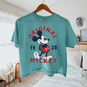 May include: A teal short-sleeve t-shirt featuring a graphic of Mickey Mouse in red shorts, with the red text "ORIGINAL" arched above and "MICKEY" below, and the black numbers "19" and "28" on either side of the character on a wooden hanger.