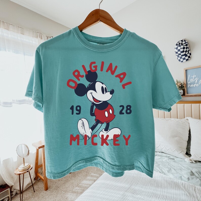 May include: A teal short-sleeve t-shirt featuring a graphic of Mickey Mouse in red shorts, with the red text "ORIGINAL" arched above and "MICKEY" below, and the black numbers "19" and "28" on either side of the character on a wooden hanger.