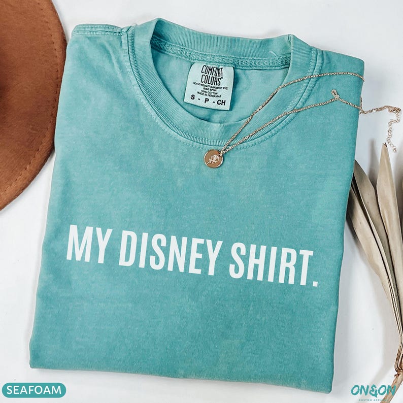 May include: Seafoam green t-shirt with the text "MY DISNEY SHIRT." A gold necklace with a pendant rests on the shirt. The shirt has a Comfort Colors tag. The shirt is folded and has a soft, worn look.