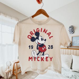 May include: A cream-colored short-sleeve t-shirt featuring a graphic of Mickey Mouse in red shorts. The text "ORIGINAL" is arched above the character, "19" and "28" are on either side, and "MICKEY" is printed in red below the character.