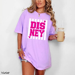 May include: A violet-colored t-shirt with the word "DISNEY" in bright pink letters, set against a white striped background. Small pink Mickey Mouse head icons are placed above and below the word. The shirt is a casual, oversized style.
