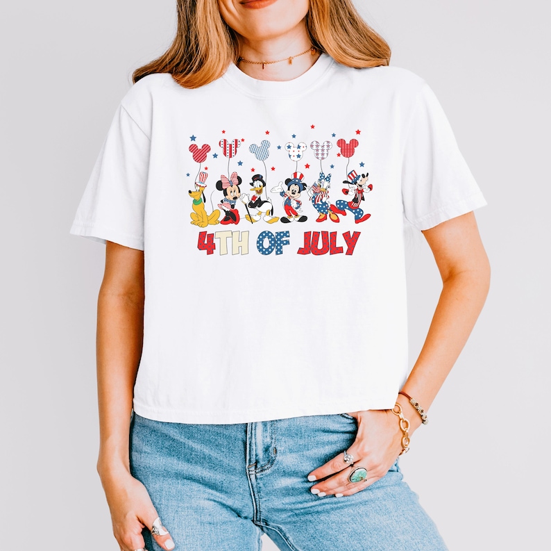 May include: A white short-sleeved t-shirt featuring a graphic of six cartoon characters holding balloons shaped like Mickey Mouse ears. The characters are dressed in red, white, and blue patriotic attire. Below the graphic is the text 4TH OF JULY.