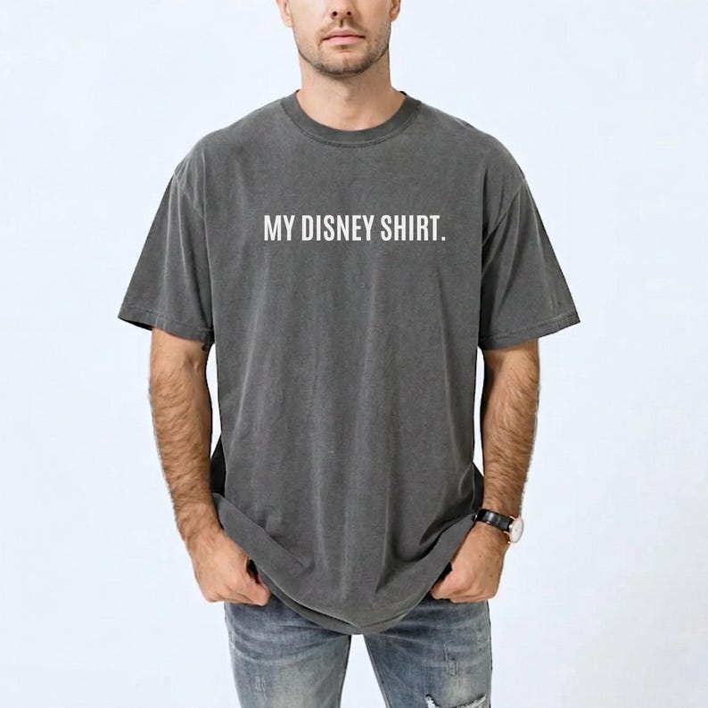 May include: Dark gray t-shirt with the text "MY DISNEY SHIRT." in white. The shirt has a slightly faded appearance and a relaxed fit. The model is wearing blue jeans and a watch.