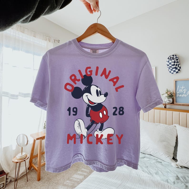 May include: A light purple short-sleeve t-shirt featuring a graphic of Mickey Mouse in the center. The text "ORIGINAL" is arched above, "19" and "28" are on the sides, and "MICKEY" is printed below in red and dark blue lettering.