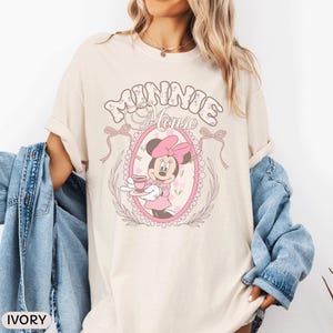 May include: An ivory short-sleeve t-shirt featuring a graphic of Minnie Mouse holding a teacup inside a pink oval frame. The text "MINNIE MOUSE" appears above the frame, which is decorated with bows and floral accents. A blue denim jacket is draped over the arms.