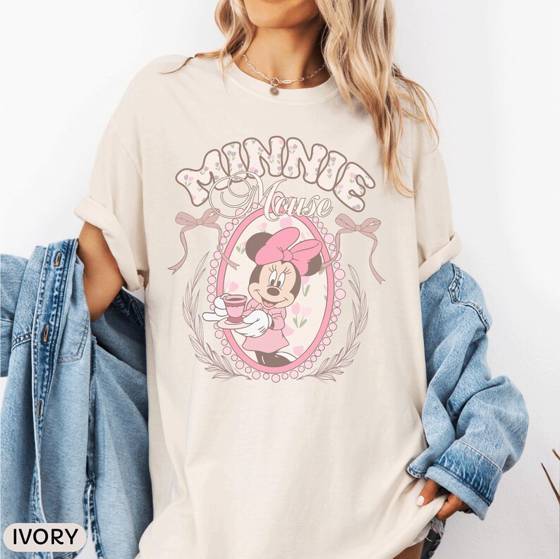 May include: An ivory short-sleeve t-shirt featuring a graphic of Minnie Mouse holding a teacup inside a pink oval frame. The text "MINNIE MOUSE" appears above the frame, which is decorated with bows and floral accents. A blue denim jacket is draped over the arms.