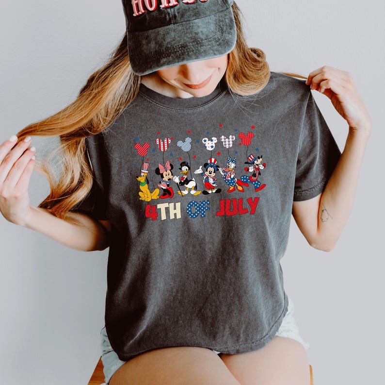 May include: A charcoal gray t-shirt featuring a graphic of Disney characters including Pluto, Minnie Mouse, Donald Duck, Mickey Mouse, Daisy Duck, and Goofy dressed in patriotic attire, holding balloons, with the text 4TH OF JULY in red and blue.