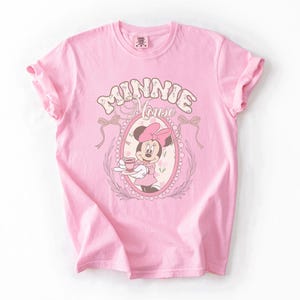 May include: A light pink short-sleeved t-shirt featuring a graphic of Minnie Mouse holding a teacup inside an oval frame. The text "MINNIE MOUSE" is printed in a stylized font above the frame, which is decorated with bows and floral accents.