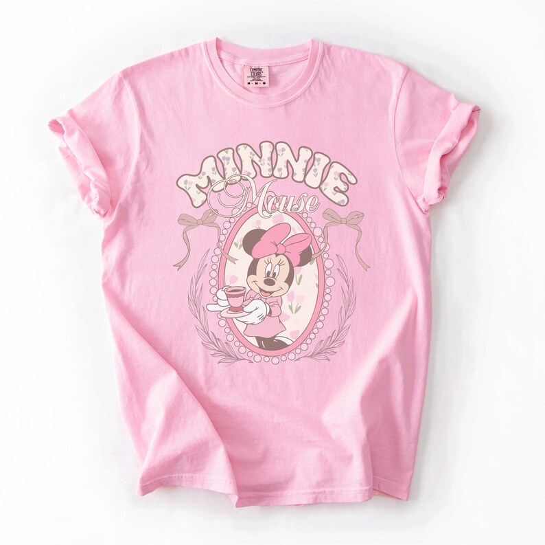 May include: A light pink short-sleeved t-shirt featuring a graphic of Minnie Mouse holding a teacup inside an oval frame. The text "MINNIE MOUSE" is printed in a stylized font above the frame, which is decorated with bows and floral accents.