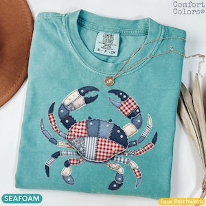 May include: Seafoam green Comfort Colors t-shirt with a faux patchwork crab design. The crab features red, white, and blue patterns, and is embellished with small white beads. A gold necklace with a pendant rests on the shirt.