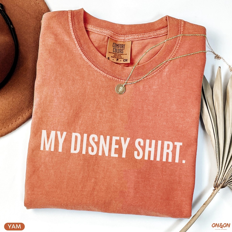 May include: Coral-colored t-shirt with the text "MY DISNEY SHIRT." A gold necklace with a pendant rests on the shirt. The shirt is folded and placed on a white surface, with a brown hat and dried plant accents.
