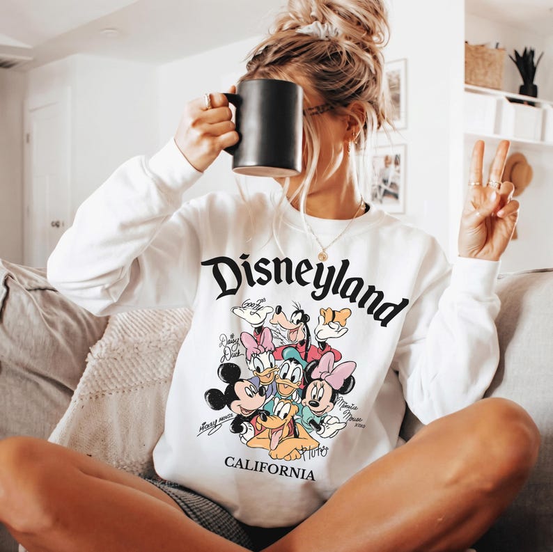 May include: White long-sleeved sweatshirt featuring the word "Disneyland" in black script, with a graphic of Disney characters below. The word "California" is printed below the graphic. A black mug is held up to the face.