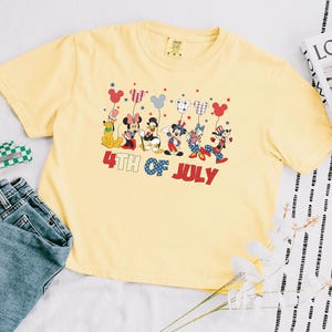 May include: A pale yellow t-shirt featuring a graphic of Disney characters including Mickey, Minnie, Donald, Daisy, Goofy, and Pluto holding balloons. The text 4TH OF JULY is printed in red and blue below the characters. The shirt has a crew neck.