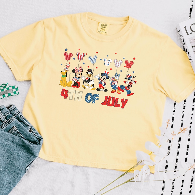 May include: A pale yellow t-shirt featuring a graphic of Disney characters including Mickey, Minnie, Donald, Daisy, Goofy, and Pluto holding balloons. The text 4TH OF JULY is printed in red and blue below the characters. The shirt has a crew neck.