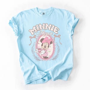 May include: A light blue short-sleeve t-shirt featuring a graphic of Minnie Mouse holding a teacup inside a pink oval frame. The text "MINNIE MOUSE" is printed in a large, decorative font above the frame, which is accented by two bows.
