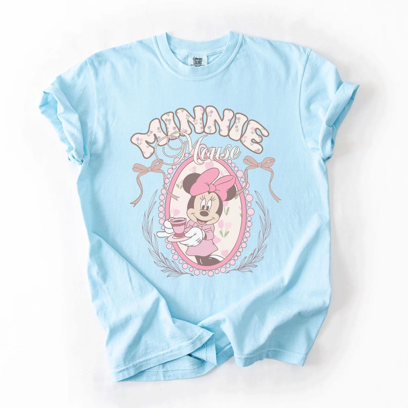 May include: A light blue short-sleeve t-shirt featuring a graphic of Minnie Mouse holding a teacup inside a pink oval frame. The text "MINNIE MOUSE" is printed in a large, decorative font above the frame, which is accented by two bows.