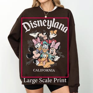 May include: A brown sweatshirt featuring a large graphic print of Disneyland characters, including Mickey Mouse, Minnie Mouse, Donald Duck, and Goofy. The word "Disneyland" is arched across the top, with "California" below. The words "Large Scale Print" are at the bottom.