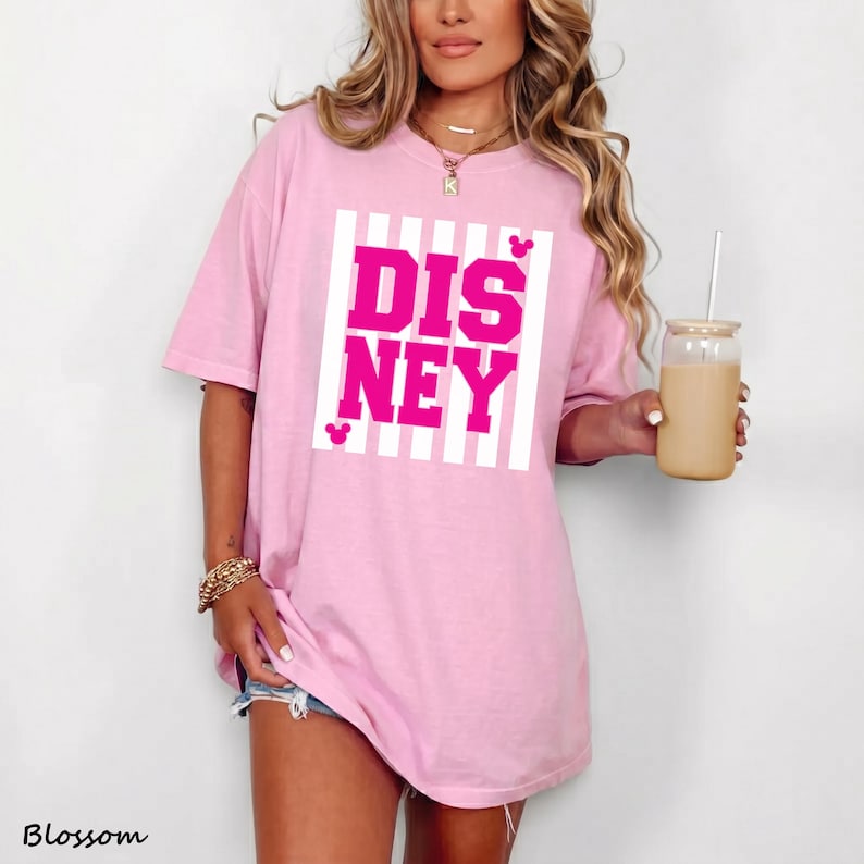 K&ouml;nnte beinhalten: Hellrosa T-Shirt mit dem Wort "DISNEY" in leuchtend pinkfarbenen Blockbuchstaben, vor einem wei&szlig; gestreiften Hintergrund. Das Shirt hat kurze &Auml;rmel und eine lockere Passform. Ein Glas mit Strohhalm wird in der Hand gehalten.