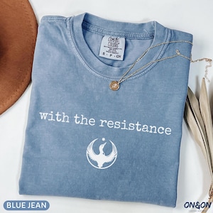 May include: A blue jean colored t-shirt with the text "with the resistance" in white. Below the text is a white graphic of a bird. A gold necklace with a pendant is draped across the shirt.