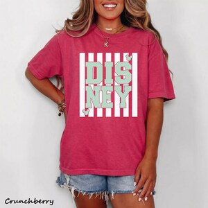 May include: A short-sleeved, cranberry-colored t-shirt with the word "DISNEY" in mint green block letters, framed by white vertical stripes. The shirt features a relaxed fit and is paired with denim shorts. The word "Crunchberry" is printed at the bottom.