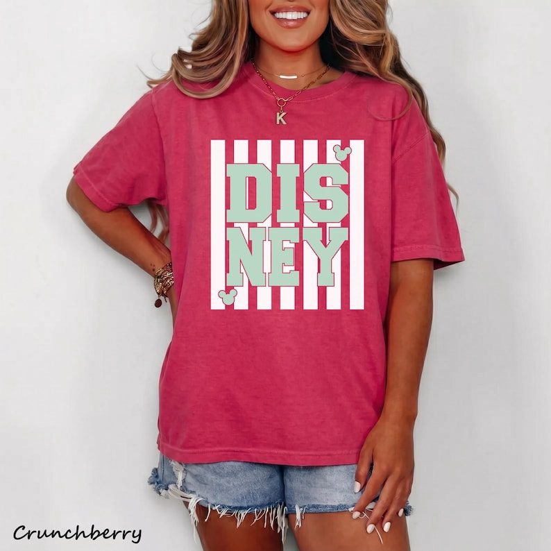 May include: A short-sleeved, cranberry-colored t-shirt with the word "DISNEY" in mint green block letters, framed by white vertical stripes. The shirt features a relaxed fit and is paired with denim shorts. The word "Crunchberry" is printed at the bottom.