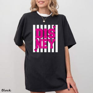 May include: Black t-shirt with the word "DISNEY" in bright pink block letters, set against a white and black striped background. Small pink hearts are placed around the word. The word "Black" is printed at the bottom left.